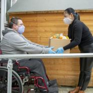 lady with a food produce delivery to the front door of a male in a wheelchair. Both people wearing face coverings.