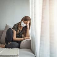 Depressed young woman sitting on couch, wearing face mask, head drooping